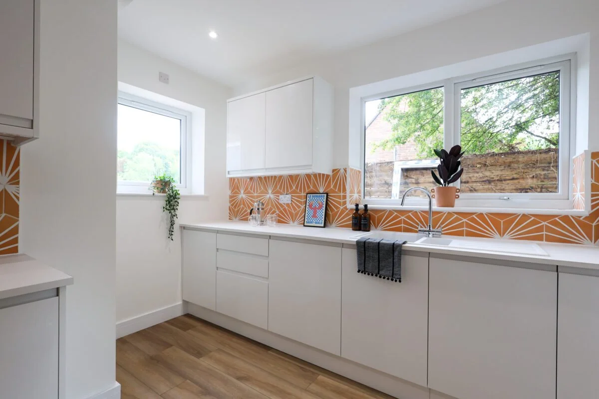 Modern white kitchen with two large windows, a white countertop, and orange geometric tile backsplash. There are small decorative items and plants on the counter, and a black and gray towel hanging from the sink.