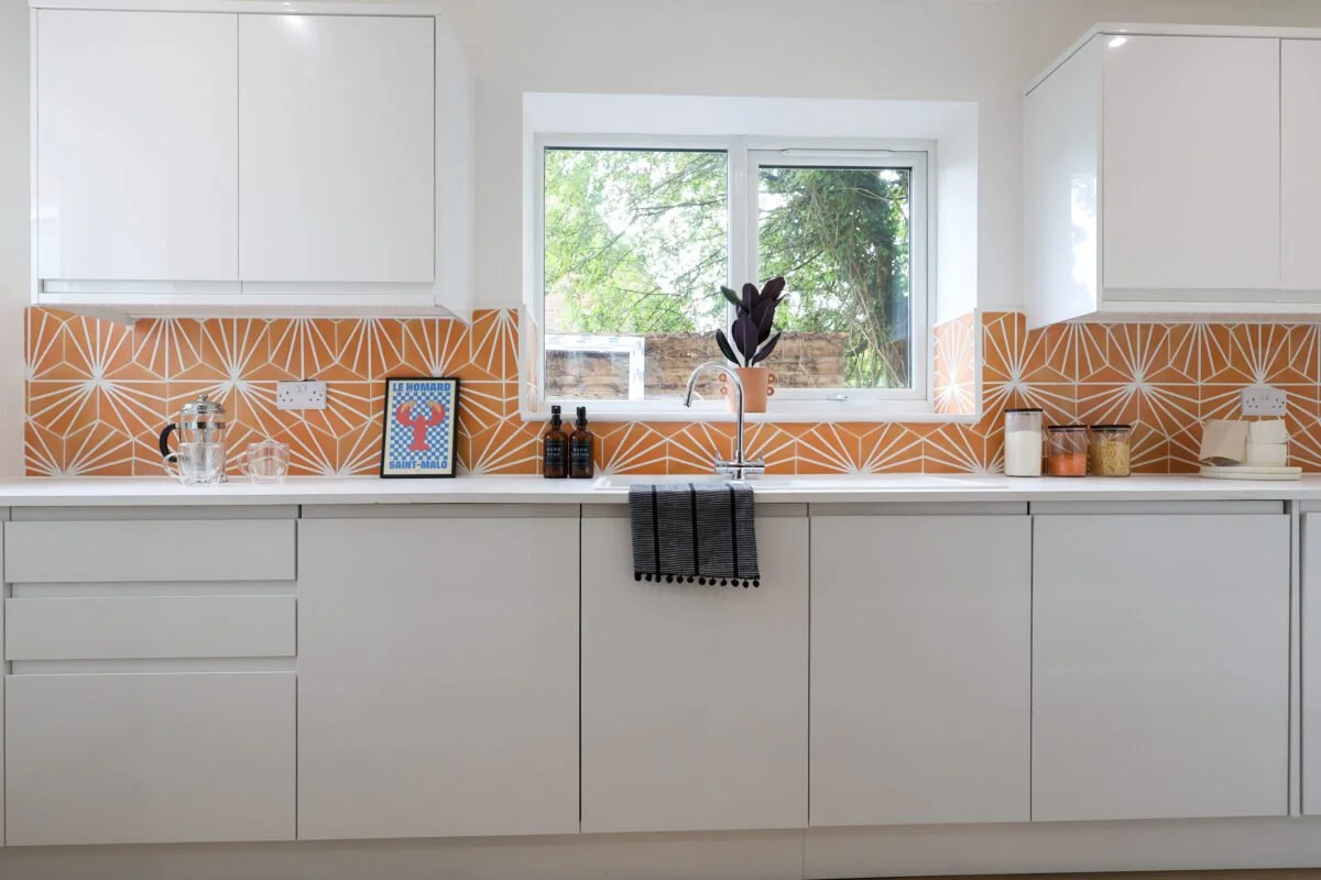 Modern kitchen with white cabinets, patterned orange backsplash, front window, and countertop with a framed poster, bottles, jars, and a plant.