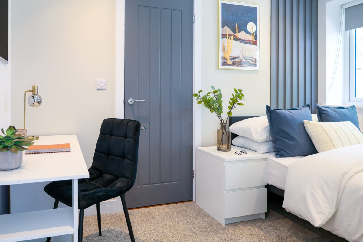 A cozy bedroom with a white bed, blue pillows, and a nightstand with a glass vase of green plants. There's a small white desk with a black cushioned chair, a lamp, and a notebook. The room has beige carpet, curtains, and a framed art print on the wal