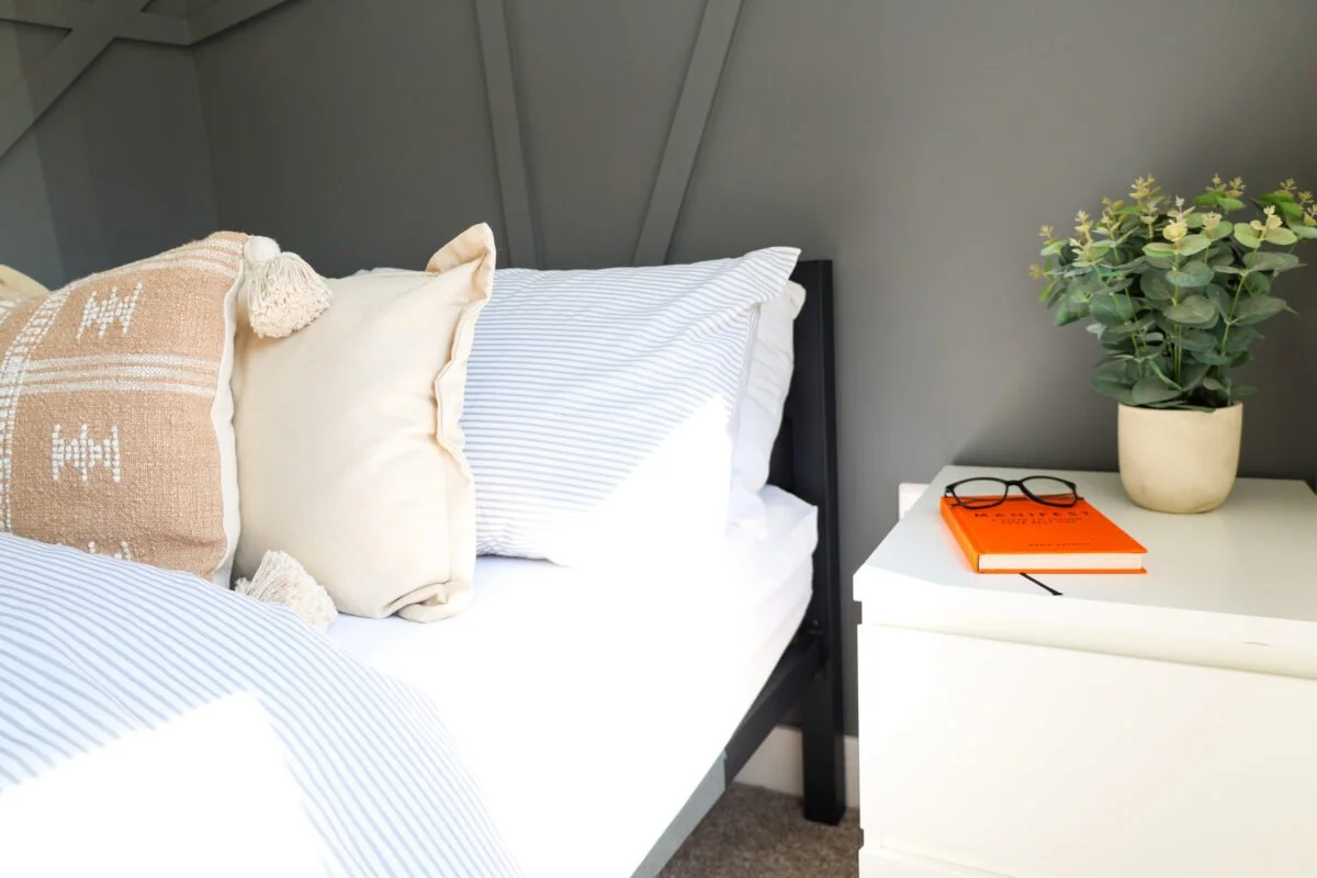 A neatly made bed with striped sheets and multiple pillows, next to a white nightstand with a potted plant, a pair of glasses, and an orange notebook.