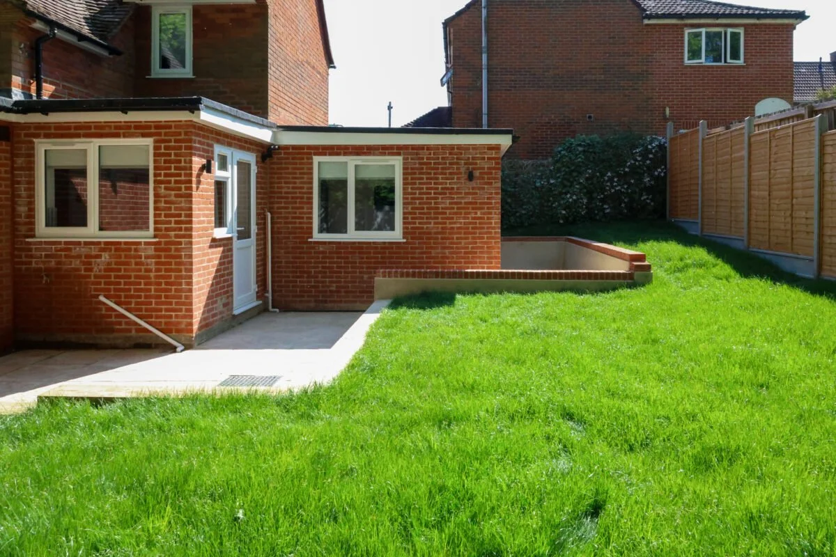 Backyard with green grass, brick house, small patio area, and wooden fence.