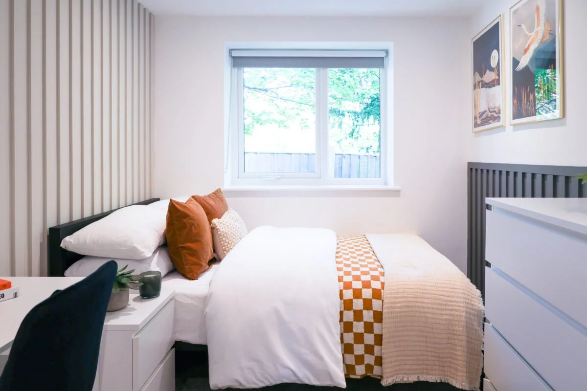 A cozy bedroom featuring a bed with white sheets, brown and cream pillows, and a beige and orange checkered blanket. There is a white nightstand with a plant and a candle, a black chair, and a dresser. The room has a window with a view of green trees
