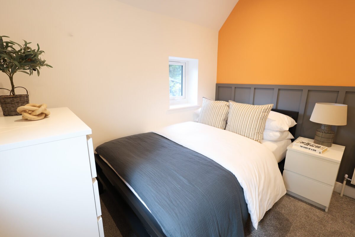 A cozy bedroom with a bed, striped pillows, and a nightstand with a lamp, window, and an orange accent wall.
