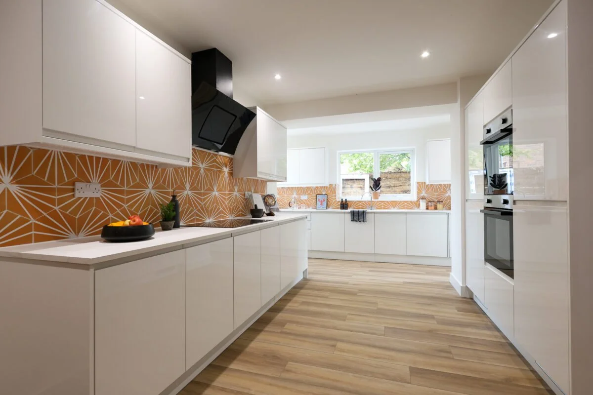 Modern kitchen with white cabinets, orange geometric backsplash, wooden floors, and a window with outdoor view.