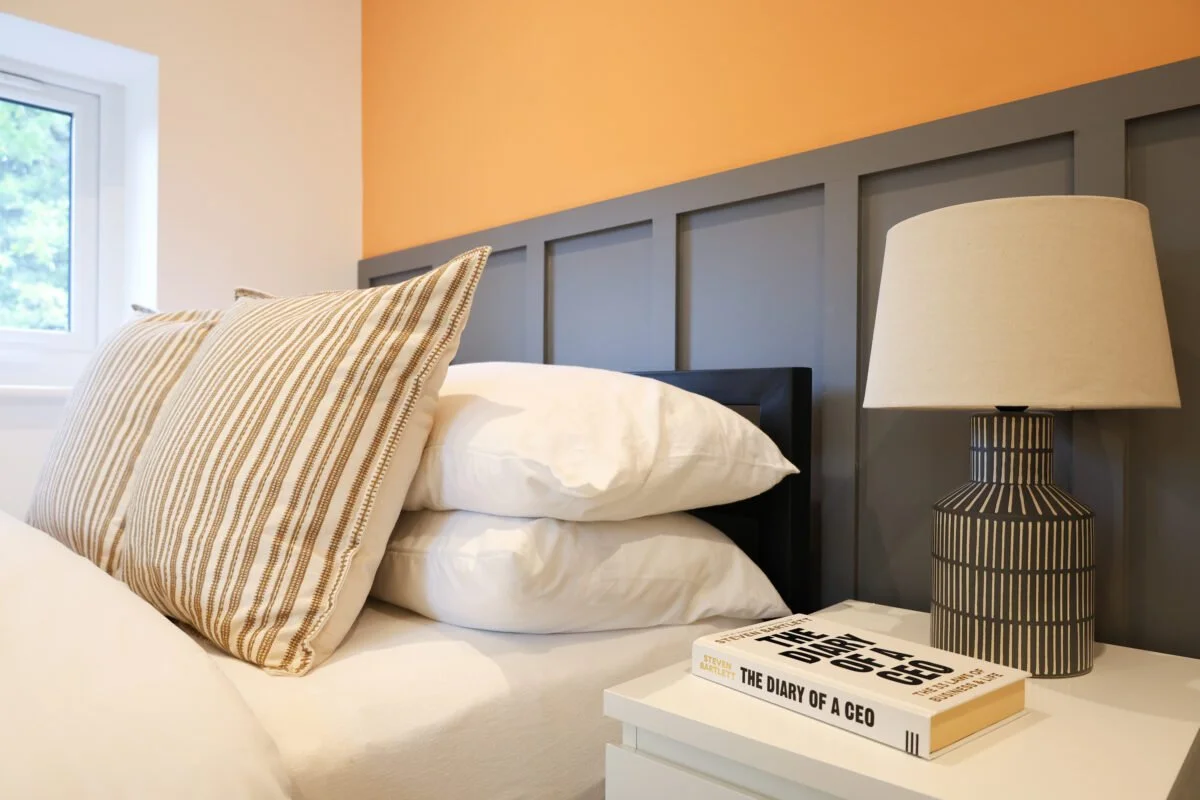 Corner of a bedroom with a bed, pillows, a nightstand, a dark striped table lamp, a book titled 'The Diary of a CEO,' and a window showing greenery outside.