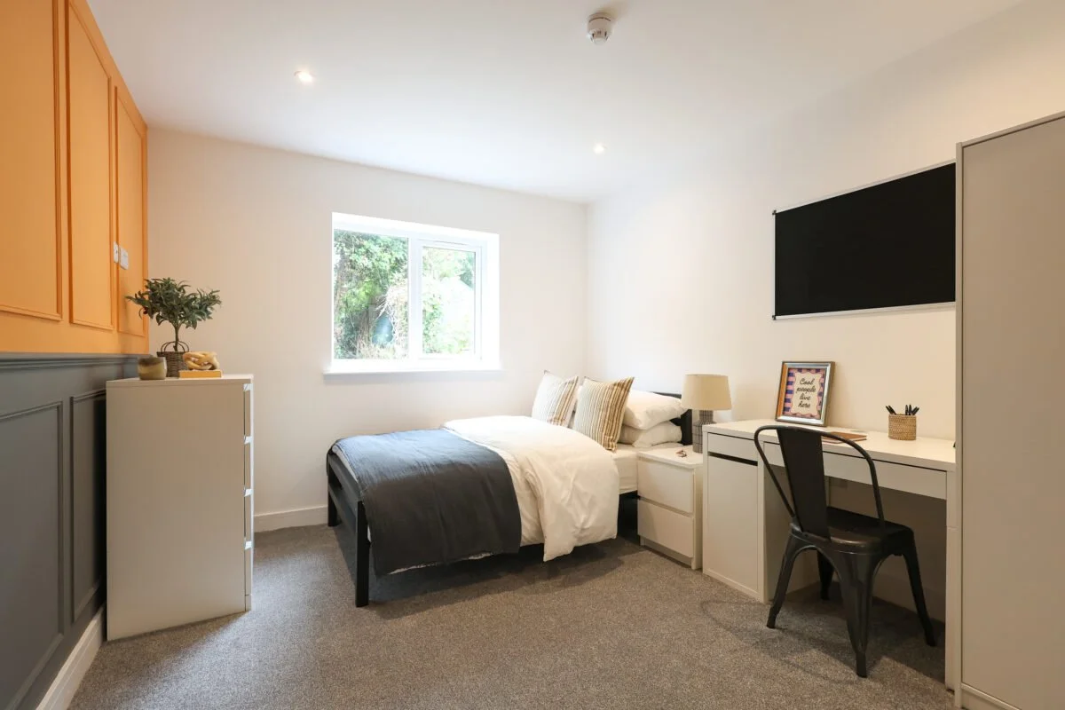 A minimalistic bedroom with a bed, white desk, black chair, dresser, and window with greenery outside.