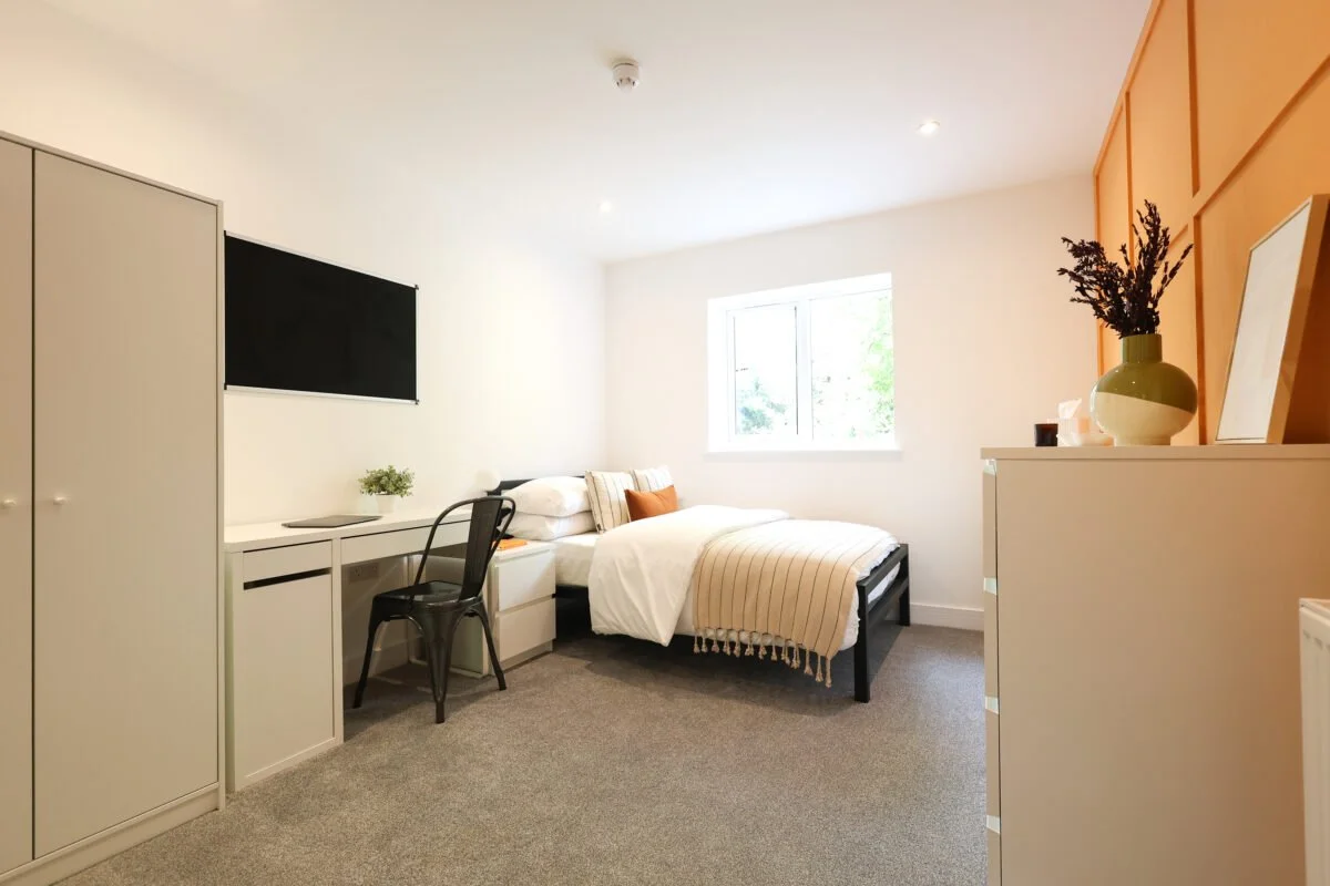 A bright, minimalistic bedroom with a bed, black desk, chair, wardrobe, and decorative items on a white dresser near a window.