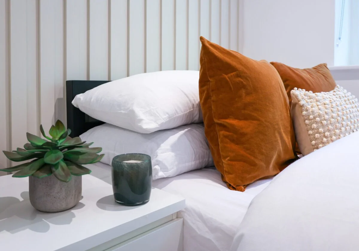 Close-up of a neatly made bed with white bedding, topped with three orange decorative pillows and one textured beige pillow. A small white nightstand next to the bed holds a potted succulent and a dark green candle. Light-colored vertical wood paneli