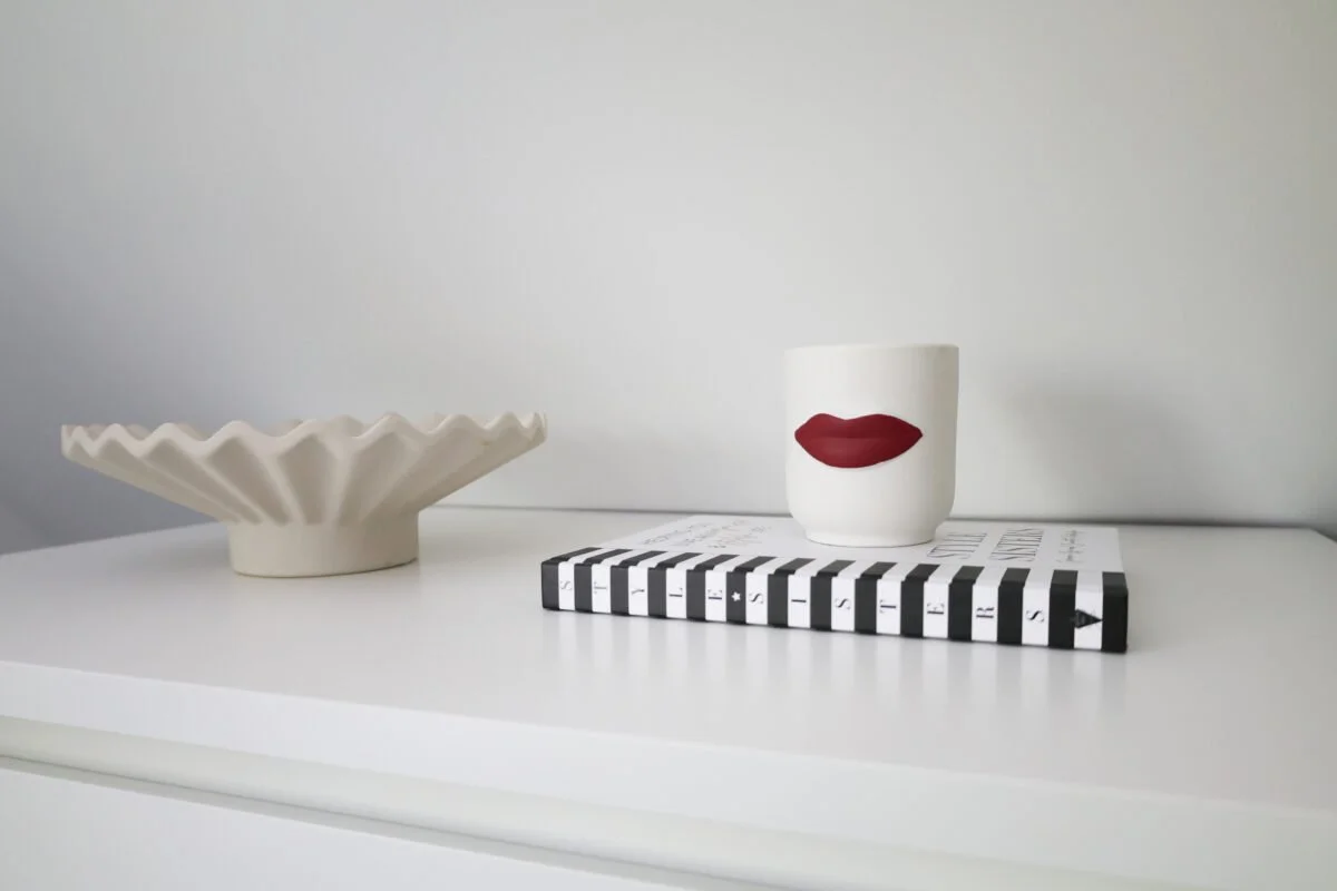 Decorative white ceramic dish, white mug with red lips design, black and white striped book, all placed on a white surface against a plain light gray background.