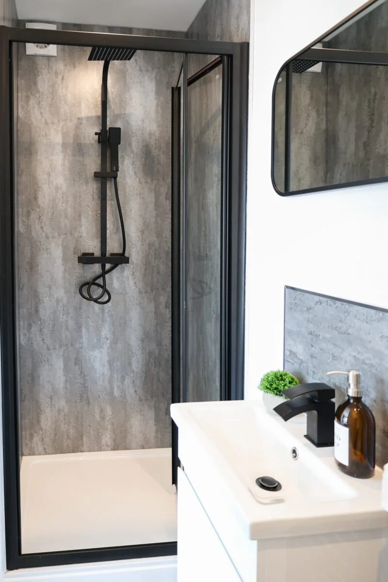 Modern bathroom with a glass-enclosed shower featuring a black showerhead and shower rod, a white vanity with a black faucet, a small plant, and a soap dispenser.