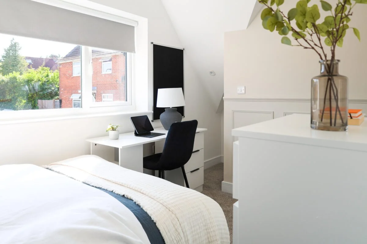 A bright bedroom with a window showing neighboring houses, a white bed with a blanket, a small desk with a laptop, a potted plant, a lamp, and a black chair, along with a vase of greenery on a white dresser.