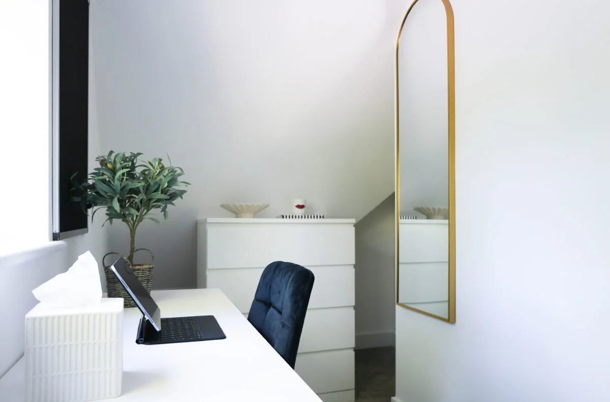 A modern workspace with a white desk, a tissue box, a tablet on a stand, a potted plant, a black chair, a white chest of drawers with decorative items, a large mirror with a gold frame, and a window with blinds.