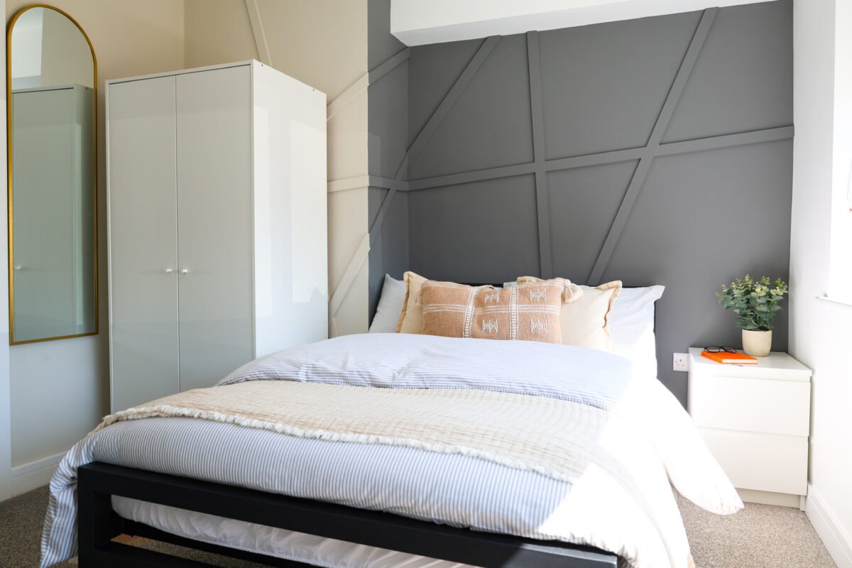 A neatly made bed with white and beige bedding, pillows, and a beige decorative pillow, in a modern bedroom with grey accent wall, white nightstand with a plant, and a wardrobe.