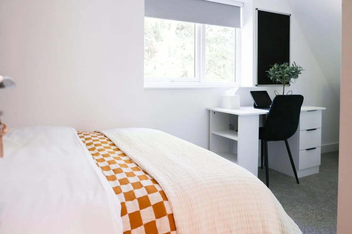 A bedroom with a white bed featuring an orange checkered blanket, a white desk with a black chair, a tablet, tissue box, and a plant near a window with white blinds.