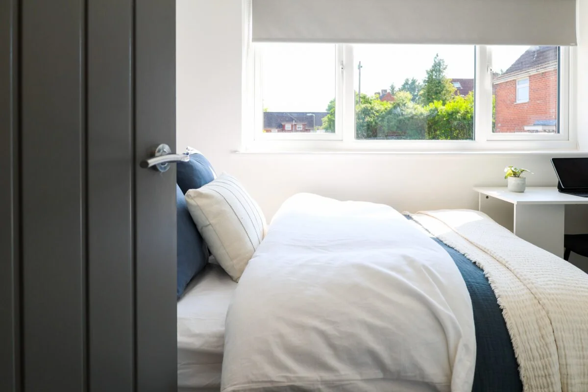 A bedroom with a bed near a large window showing green trees and houses outside, a white desk with a potted plant and a laptop, and a partially opened door.