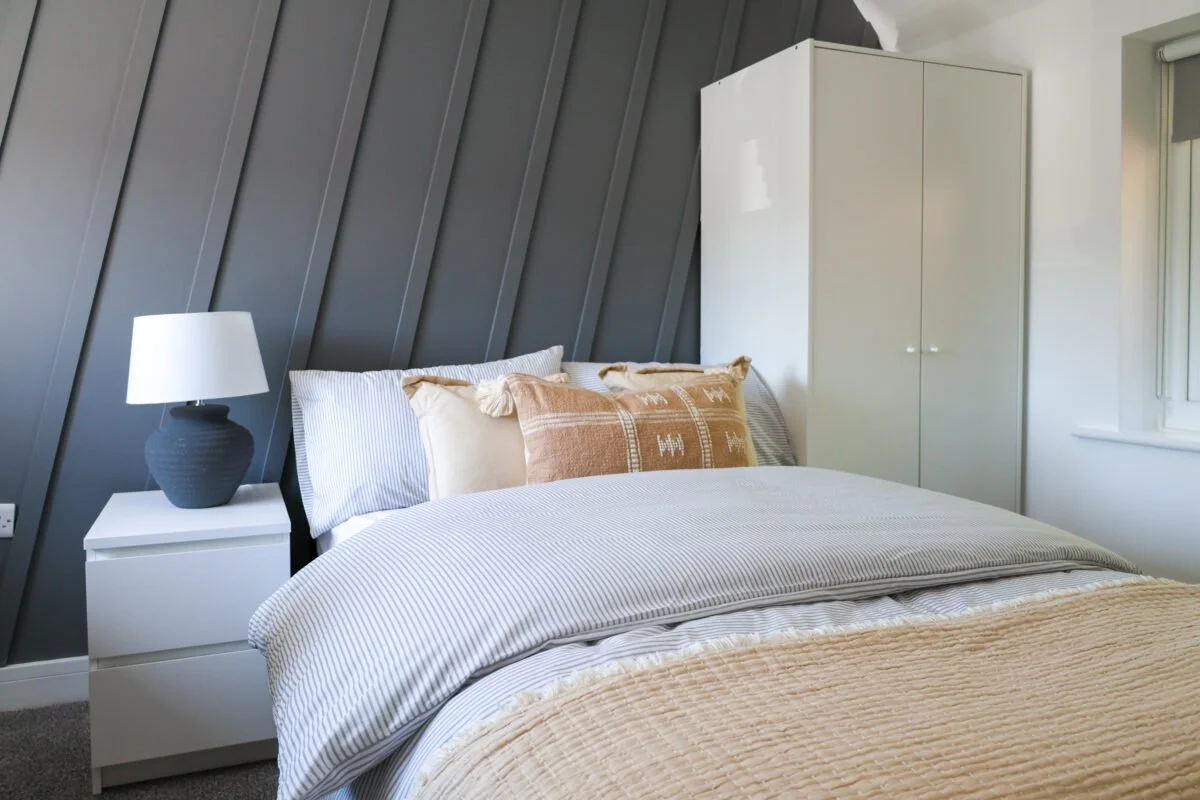 A neatly made bed with white and striped bedding, accented with beige and patterned pillows, a white nightstand with a textured dark blue lamp, and a white wardrobe against a gray, sloped wall in a bedroom with a window.