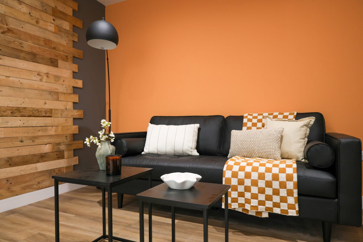 Living room with a black leather sofa, wooden wall accent, orange wall, and decorative pillows with checkered and textured patterns. There are two black coffee tables, one with a white bowl and the other with a vase of flowers, and a black floor lamp