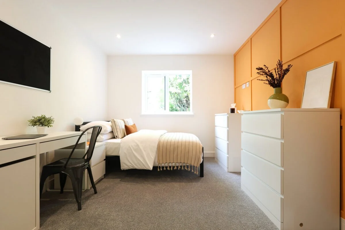 A modern bedroom with a bed, dresser, and desk, with a window showing greenery outside, decorated in neutral tones with orange accent wall.