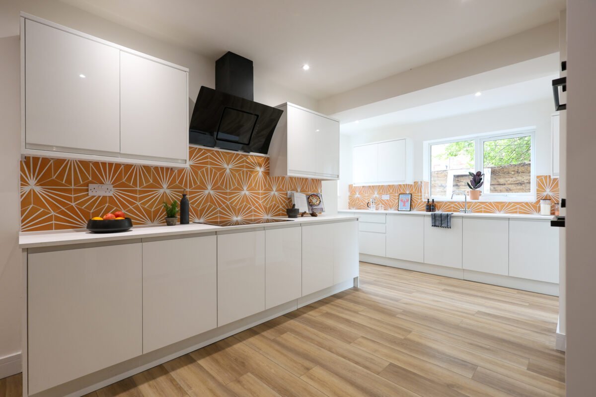 Modern kitchen with white cabinets and orange geometric backsplash, wood flooring, and a large window letting in natural light.