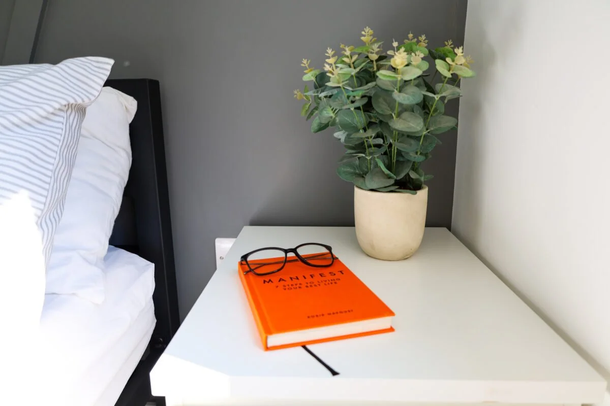 A white nightstand with an orange book titled 'MANIFEST' and a pair of black eyeglasses on top, next to a potted green plant in a beige pot, positioned beside a bed with white and gray striped sheets.