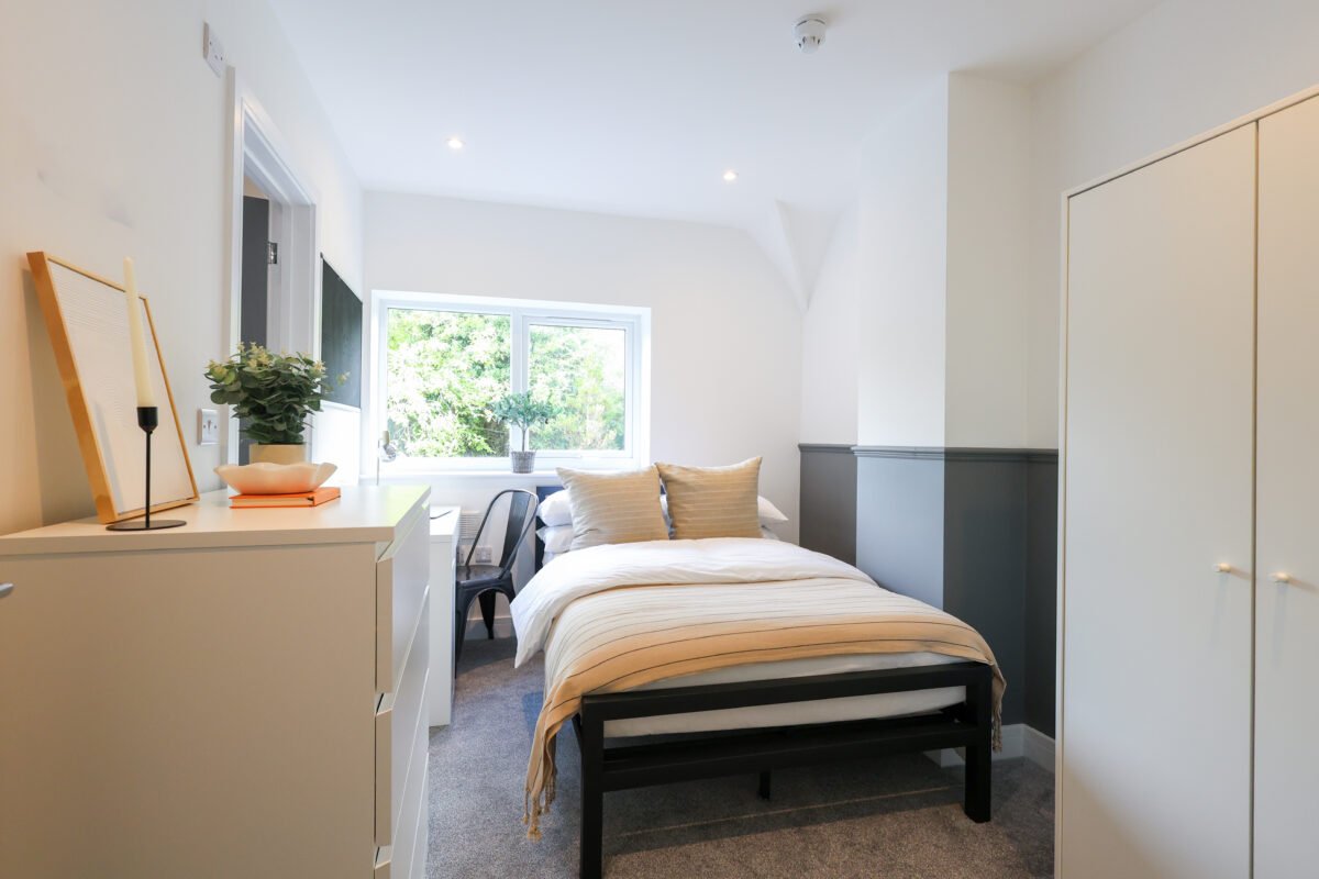 Bright bedroom with white walls, a bed with beige and white striped bedding, two beige pillows, and a window with greenery outside. There is a white dresser with decorative items, a plant, and a candle. A small black chair is next to the window.