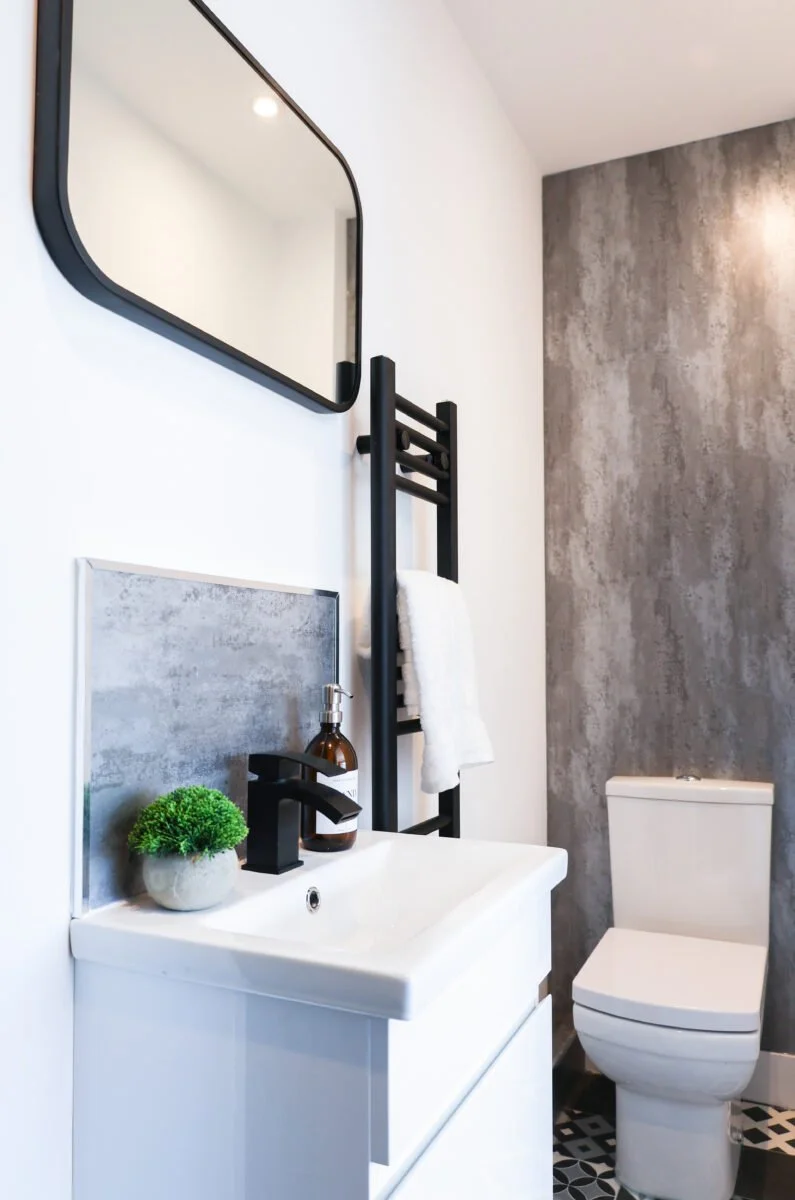 Modern bathroom with white walls, a rectangular mirror, a small white sink with a black faucet, a potted plant, a brown soap dispenser, a black towel rack with a white towel, a textured gray accent wall, and a toilet.
