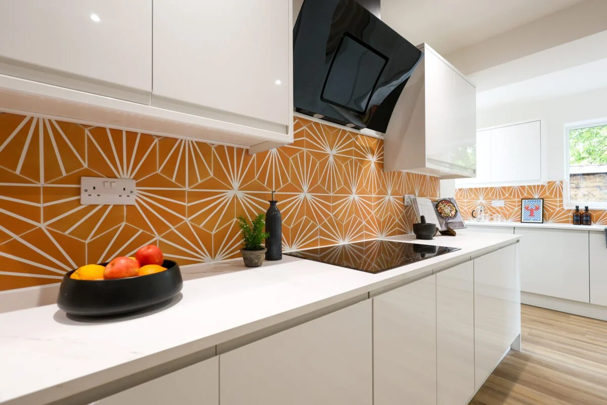 Modern kitchen with white cabinets, orange geometric patterned backsplash, black stovetop, and window with natural light.