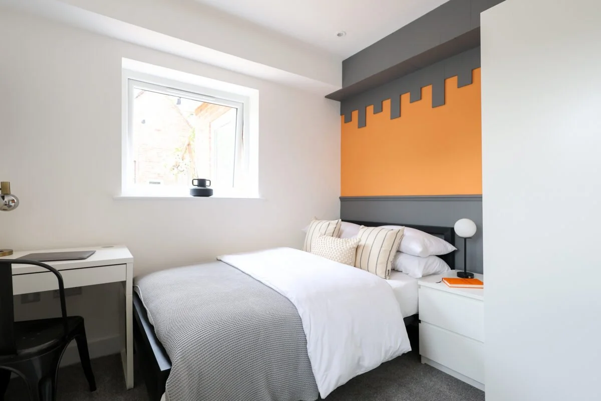Modern bedroom with a white bed, decorative pillows, a white bedside table with a lamp and orange notebook, a window with sunlight, and a wall with orange and gray geometric design.