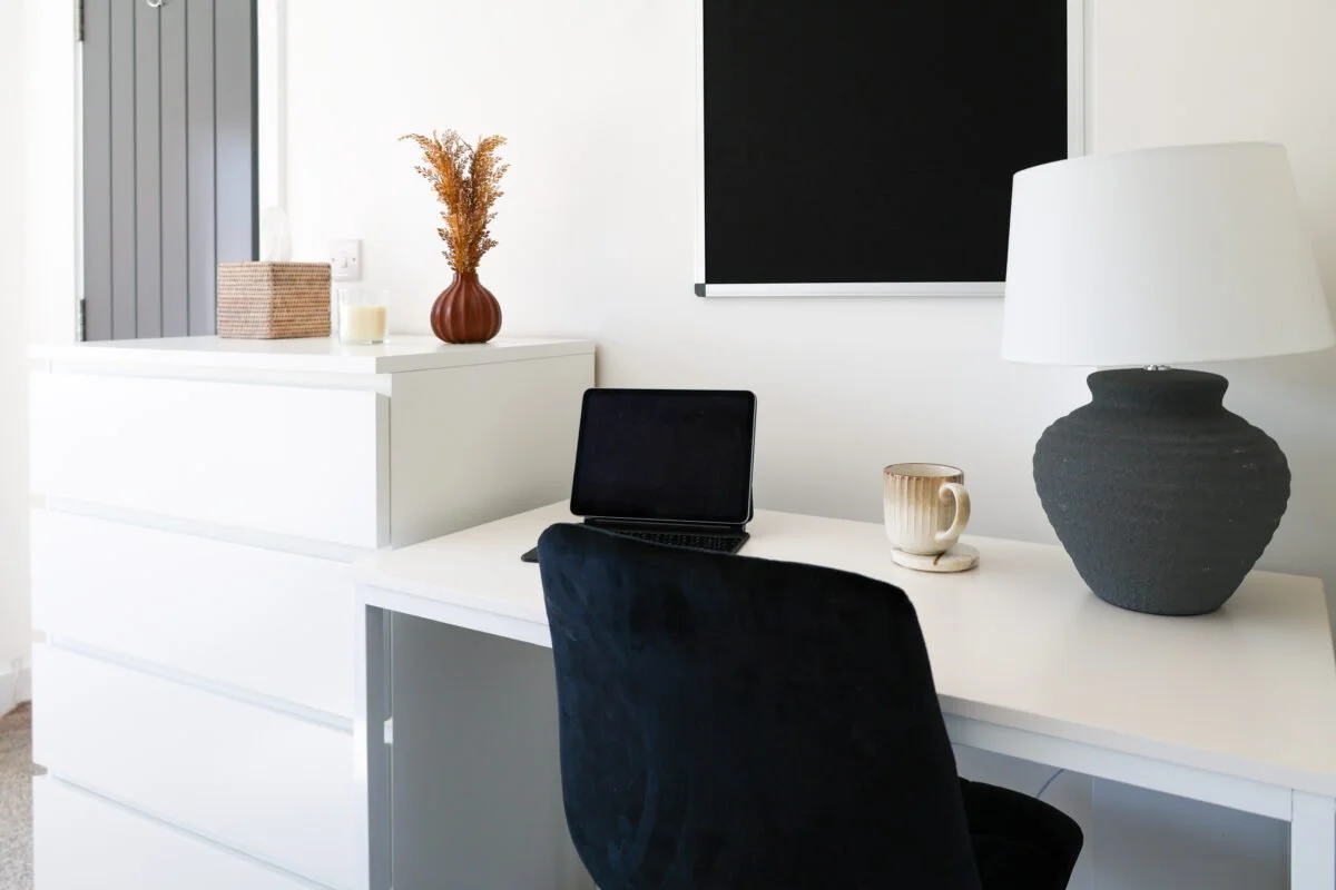 Modern home office with a white desk, a black laptop, a white cup, a large gray lamp, and decorative items including a vase with dried flowers on a white cabinet.