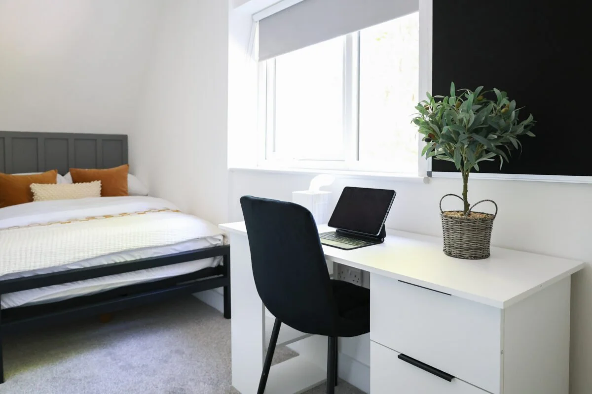 A bedroom with a bed, pillows, a window, a desk, a chair, a laptop, a tissue box, a plant in a woven basket, and a blackboard.