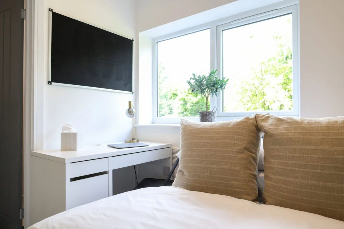 Bright bedroom with a large window, beige pillows on a bed, a white desk, a black chalkboard, a table lamp, and a potted plant.