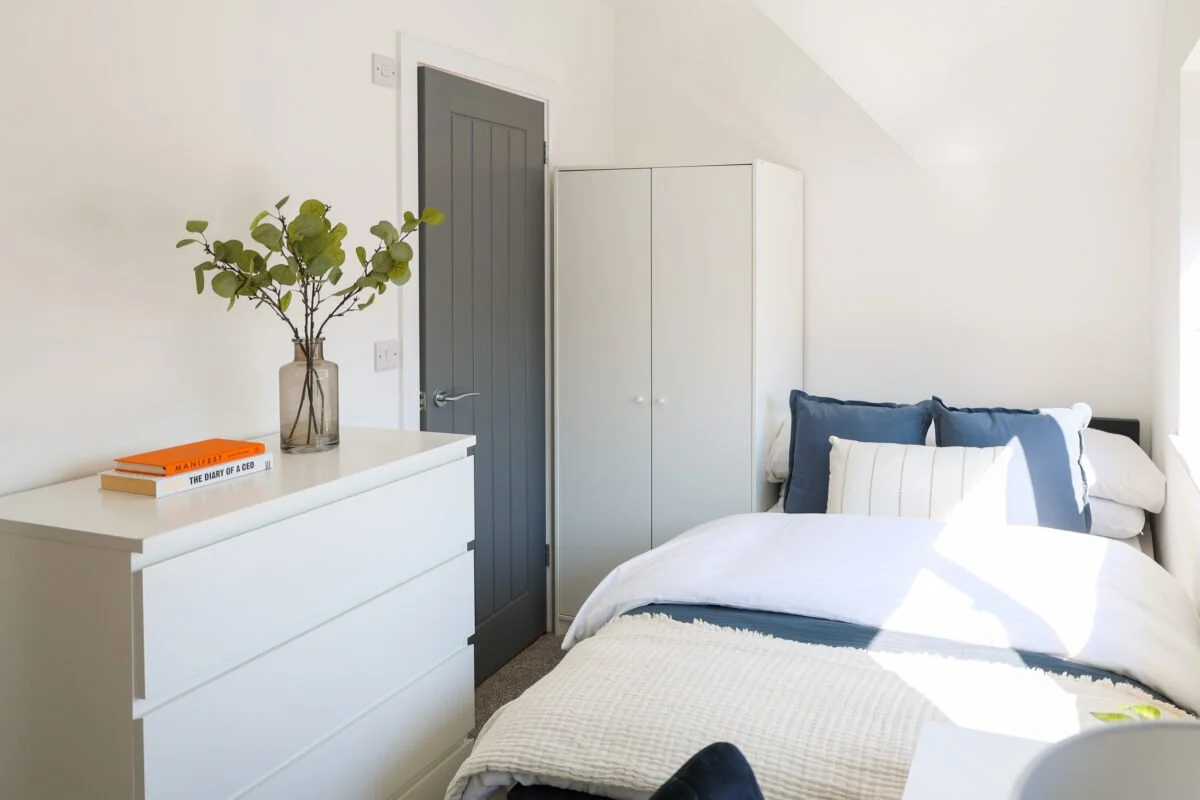 A minimalist bedroom with a white dresser topped with a vase holding green foliage and two books, one titled 'MANIFEST' and the other 'THE DIARY OF A CEO', next to a grey door and a white wardrobe, with a bed featuring white and blue pillows and natu