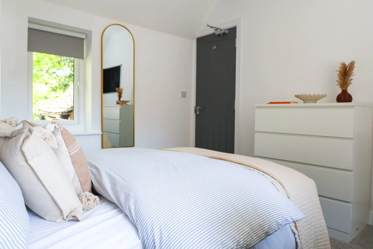 A neatly made bedroom with a bed featuring neutral-colored pillows, a window with a view of a tree, a mirror with a gold frame, a white dresser with decorative items, and a dark gray door.
