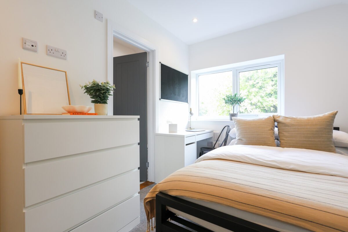 Bright bedroom with a bed, nightstand, desk, and dresser, with large window showing outside greenery.