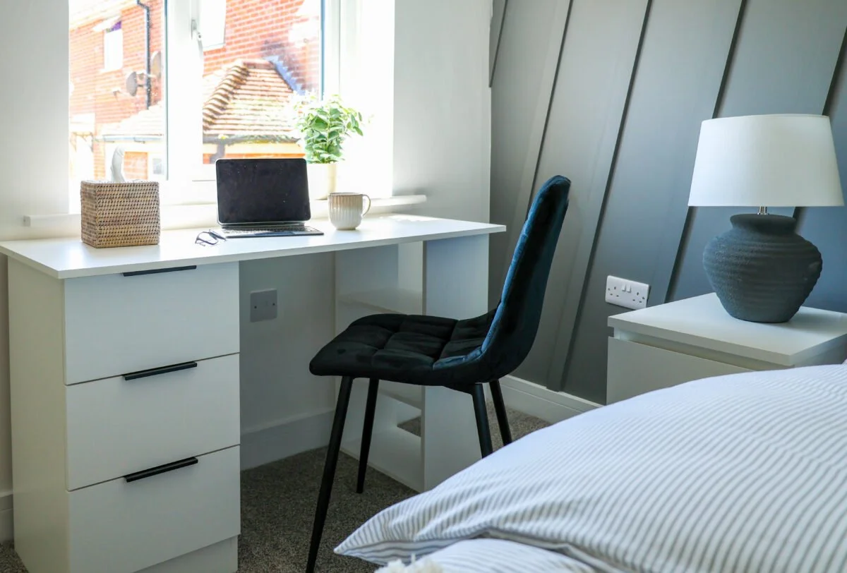 A cozy bedroom with a white desk near a window, a black chair, a laptop, a mug, a plant, a wicker basket, and a white bedside table with a gray lamp.