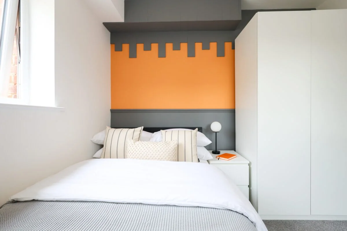 A neatly made bed with white bedding and multiple pillows, positioned in a modern bedroom with a gray and orange accent wall, a small white nightstand with a modern lamp and an orange book, and a white wardrobe on the right.