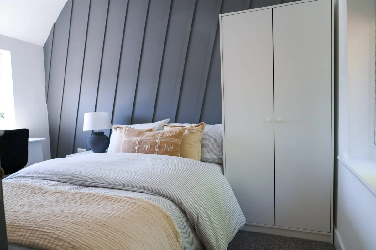 A neatly made bed with beige and striped pillows, a dark gray lamp on a nightstand, and a white wardrobe in a bedroom with sloped ceiling and gray accent wall.