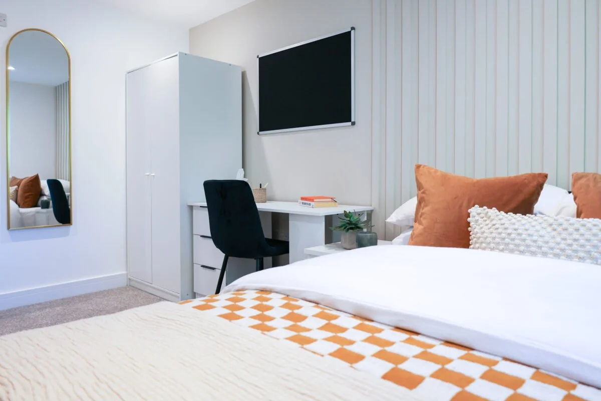 A cozy bedroom with a bed featuring white, brown, and textured pillows, a white desk with a black chair, a wardrobe, a wall-mounted blackboard, and a mirror on the wall.