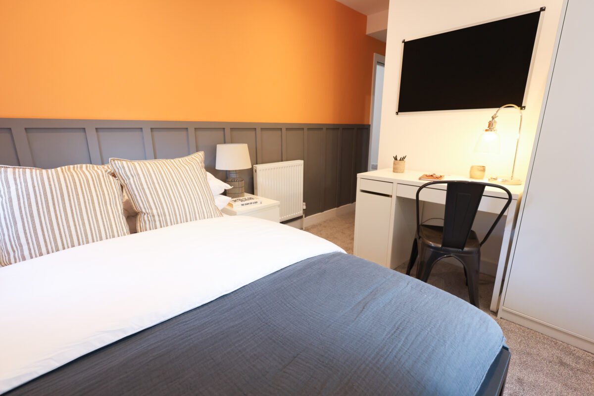 A cozy bedroom with a bed, striped pillows, a nightstand with a lamp, a small desk with a chair, and a blackboard or chalkboard on the wall.