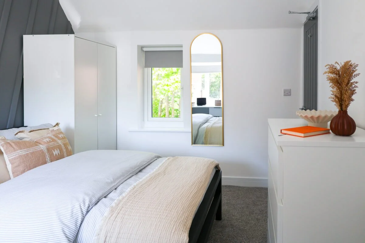 A cozy bedroom with a white bed, beige and white pillows, a white closet, a window with greenery outside, a tall mirror, and a white dresser with a brown vase containing dried flowers and an orange book.