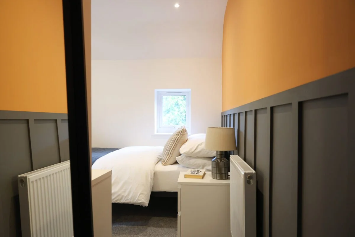 A cozy bedroom with a white bed, striped pillows, a bedside lamp, a small window, and a white nightstand with a book and telephone on it.