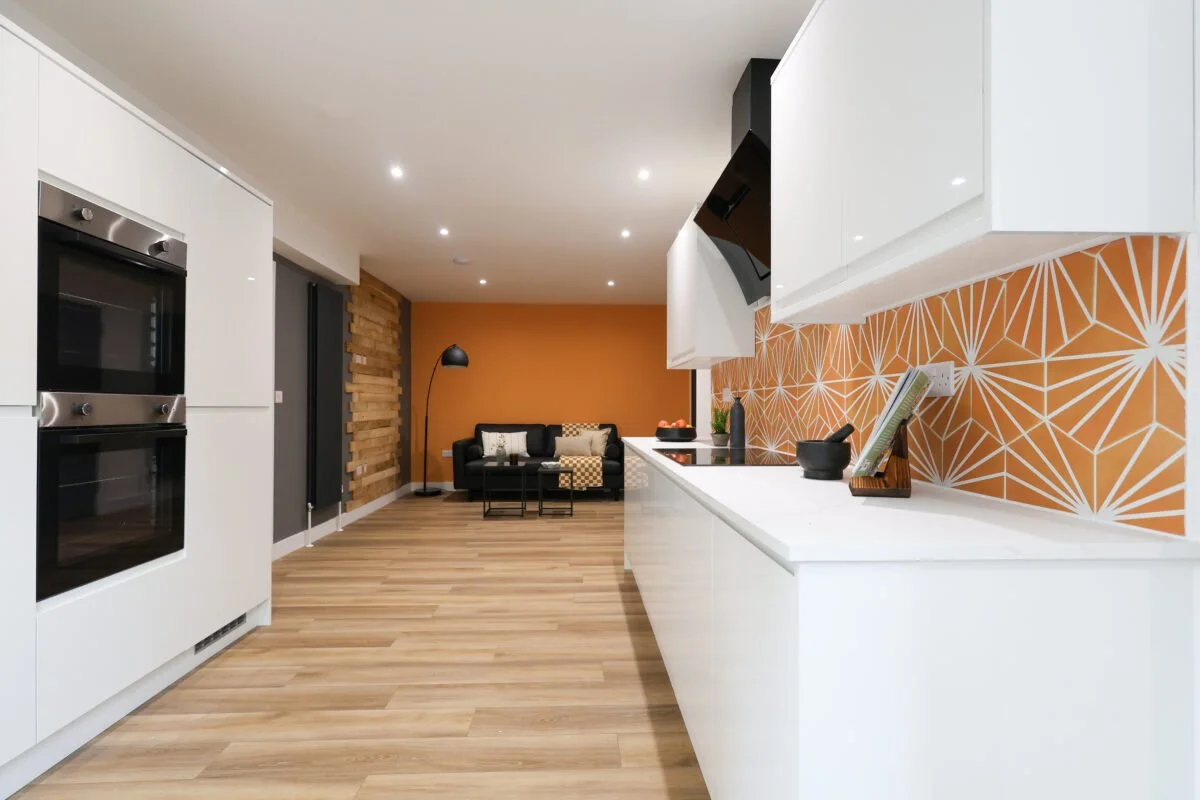 Modern open-concept kitchen with white cabinets, orange geometric backsplash, and a view into a living area with a black sofa and orange accent wall.