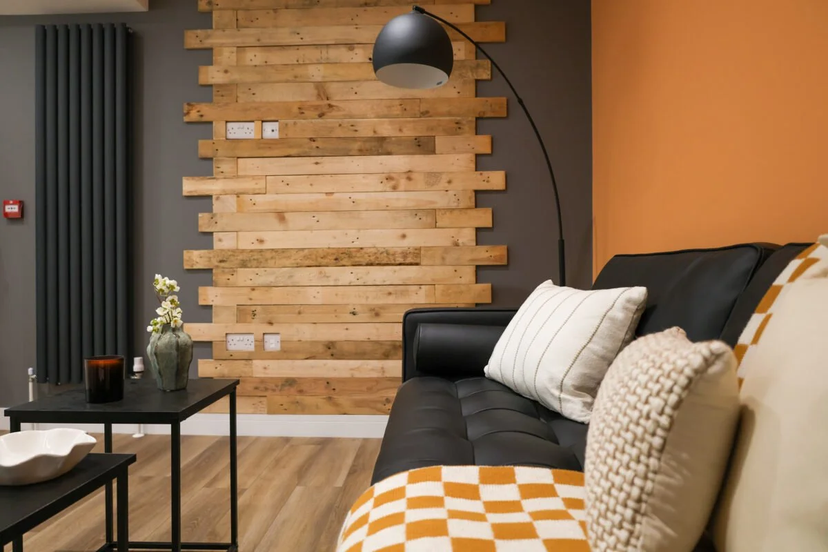 Living room with a black leather sofa, decorative pillows, wooden wall panel, black floor lamp, and a black coffee table with a vase and decorative objects.