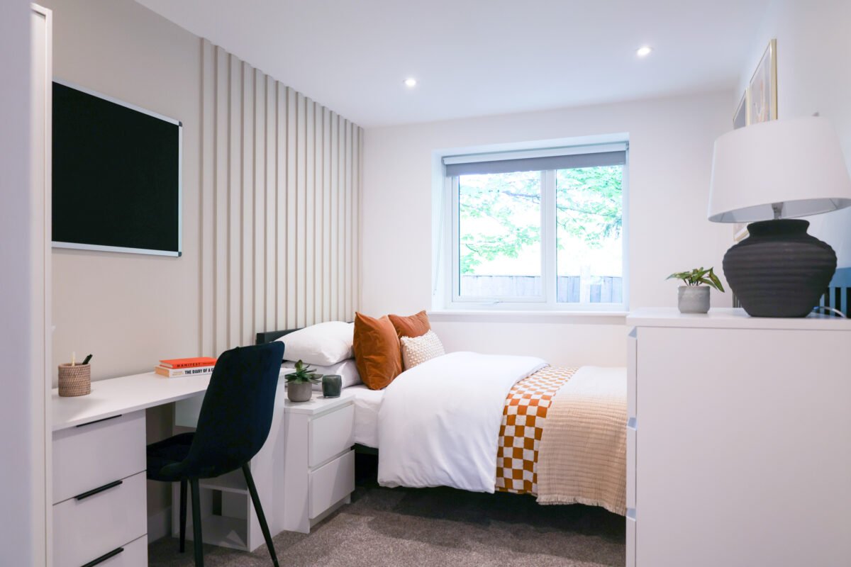 Bright, modern bedroom with a bed, decorative pillows, a window with a view of trees, a white desk with books and a chair, and a dresser with a black lamp and potted plants.