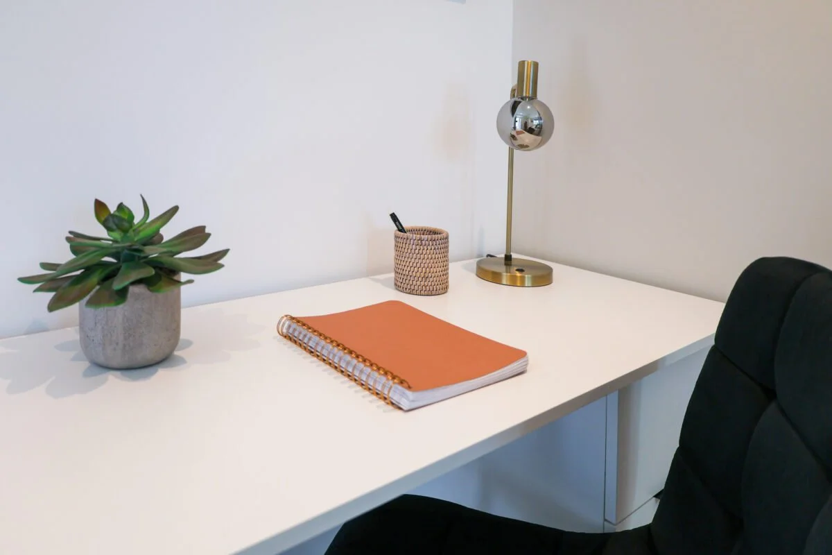 Minimalist white desk with a potted succulent, a pink spiral notebook, a small woven pen holder with pens, a gold and chrome modern desk lamp, and a black office chair.