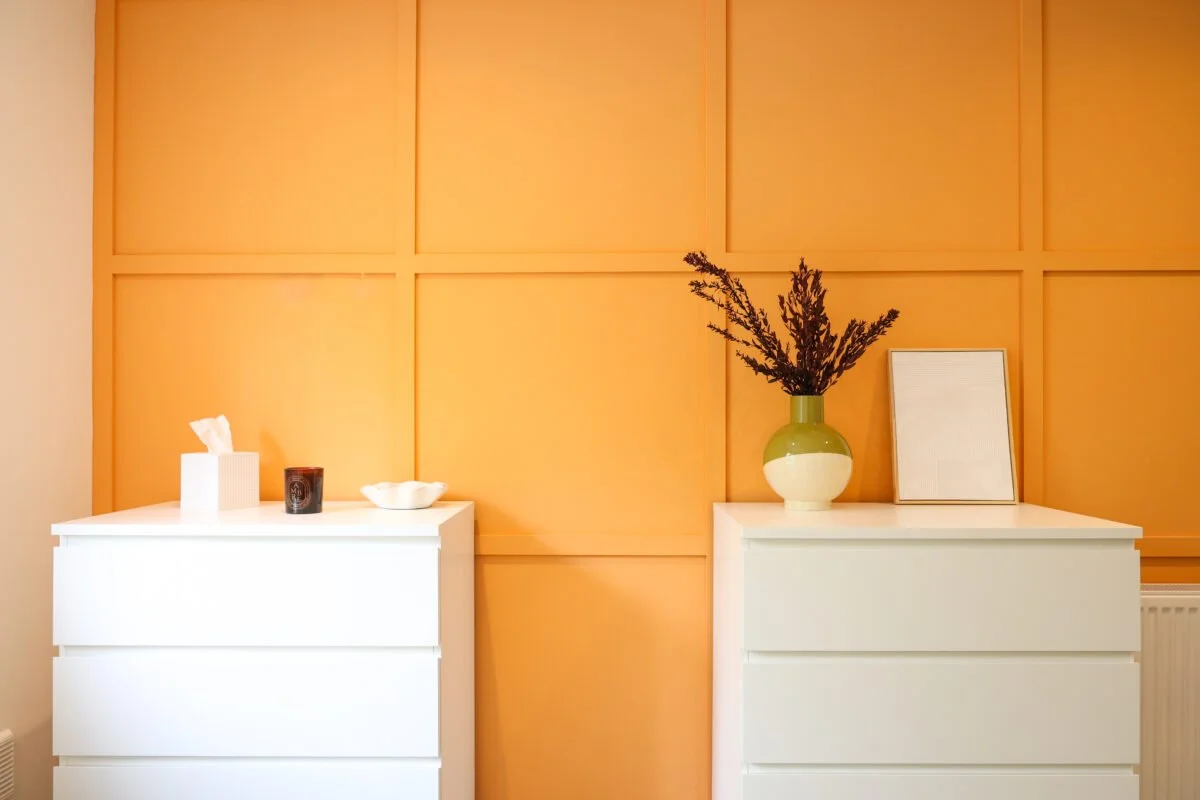 Two white dressers against a yellow paneled wall, decorated with a tissue box, black candle, white bowl, green and cream vase with dried branches, and a picture frame.