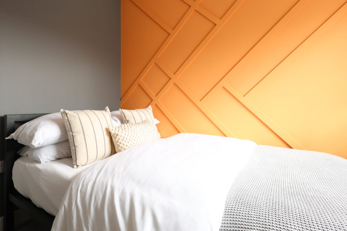 A neatly made bed with white bedding and striped pillows, set against a wall with a geometric wooden paneling design painted in orange.