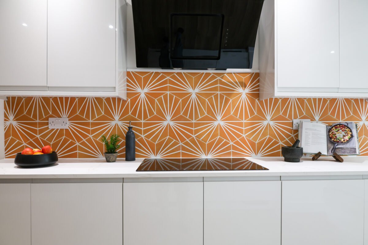 Modern kitchen with white cabinets, orange geometric backsplash, black induction cooktop, black and green decorative items, and open cookbook with food images.