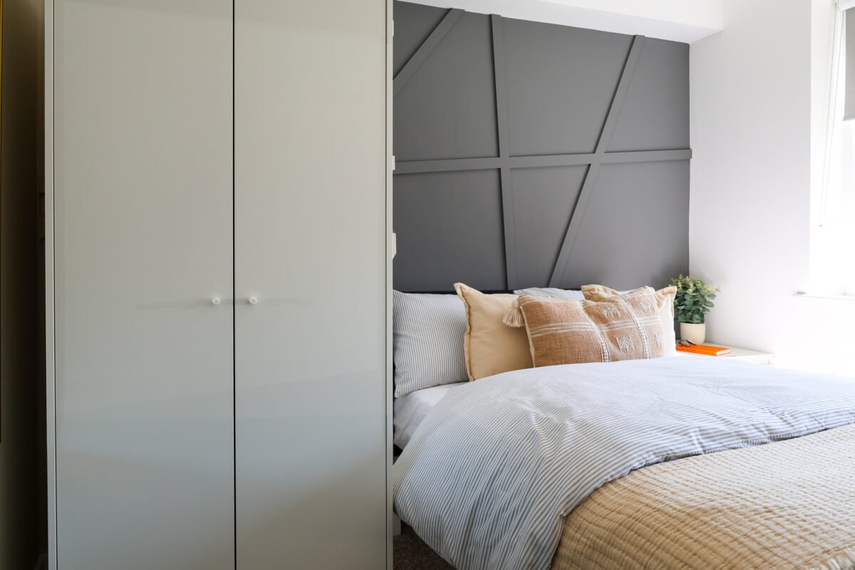 A bedroom with a bed, pillows, a gray geometric headboard, a white wardrobe, a window with white curtains, and a small bedside table with a plant, a book, and an orange object.