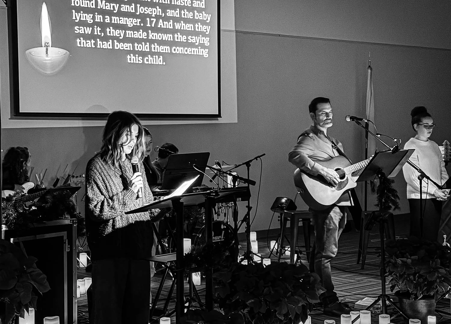 A group of musicians performing on stage with a woman holding a microphone and a man playing a guitar, with a large screen displaying text about a biblical story in the background. The stage is decorated with candles and plants.